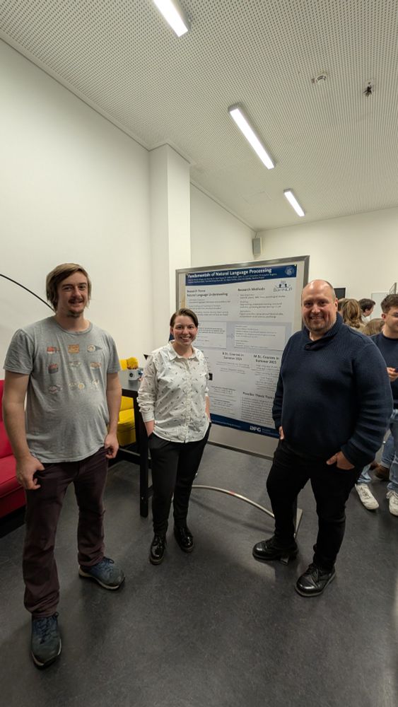 A group of three people is posing in front of a research poster titled "Fundamentals of Natural Language Processing." The poster contains sections on research focus, research methods, and possible thesis topics. The individuals are standing in an indoor academic or conference setting with other attendees visible in the background. One person is wearing a casual t-shirt with cartoon illustrations, another is in a button-up shirt, and the third is dressed in a dark sweater. The setting includes modern lighting and furniture, with a red couch and a yellow chair visible on the left side of the image.

