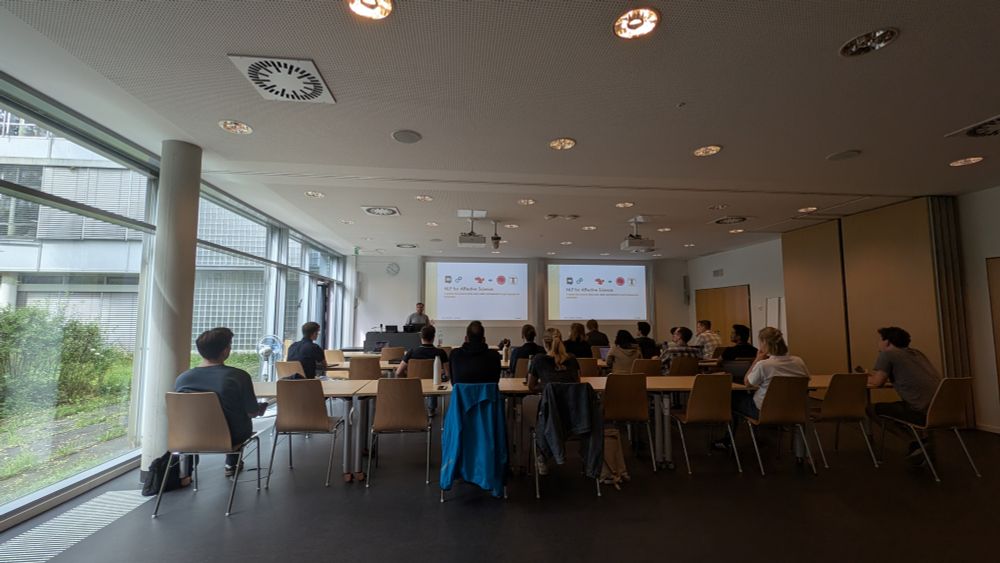 photo of a seminar room from the back to the front. many people sit at desks and listen to a presentation. in the front Saif Mohammad gives a talk. There are two projections next to each other of his slides.