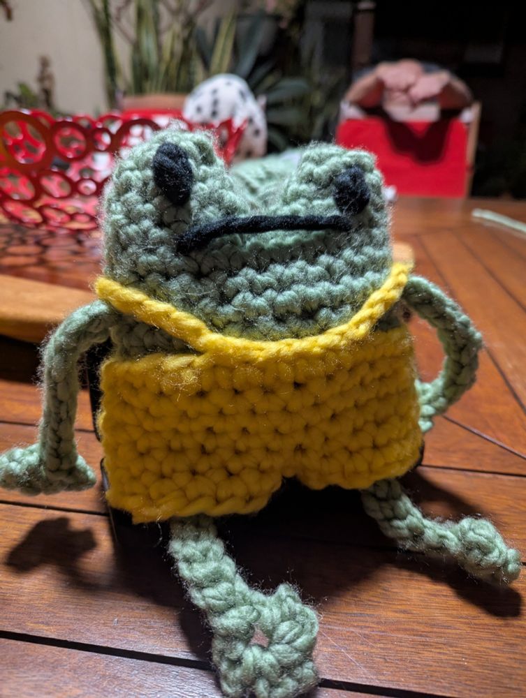 A stylised green frog wearing yellow overalls, all crocheted.