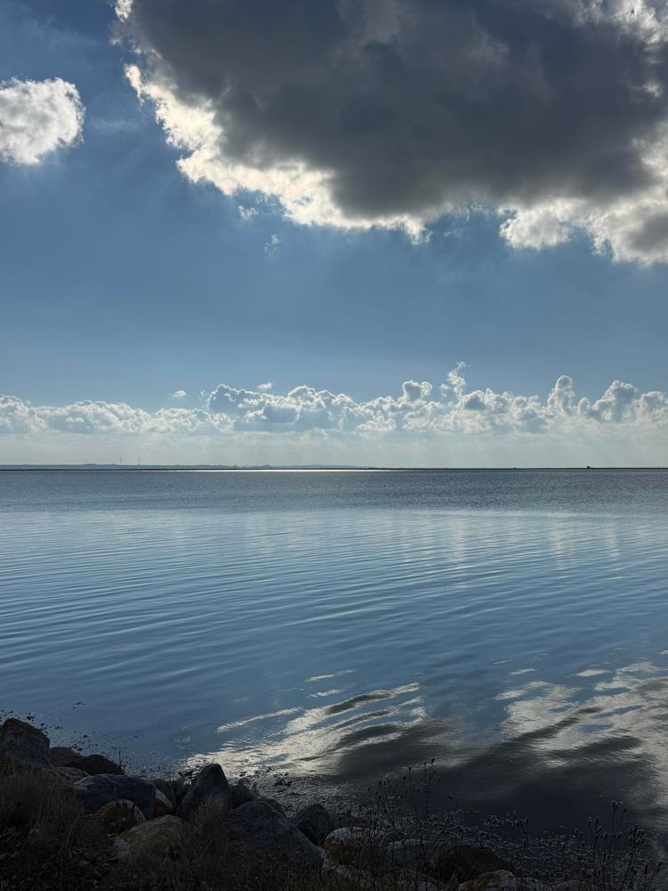 Wasser, das den Himmel reflektiert. Der Horizont zeigt eine Mischung aus Wolken und Sonnenlicht, wobei dunkle Wolken sich von helleren abheben. Felsen sind im Vordergrund sichtbar, mit trockener Vegetation in der Nähe.