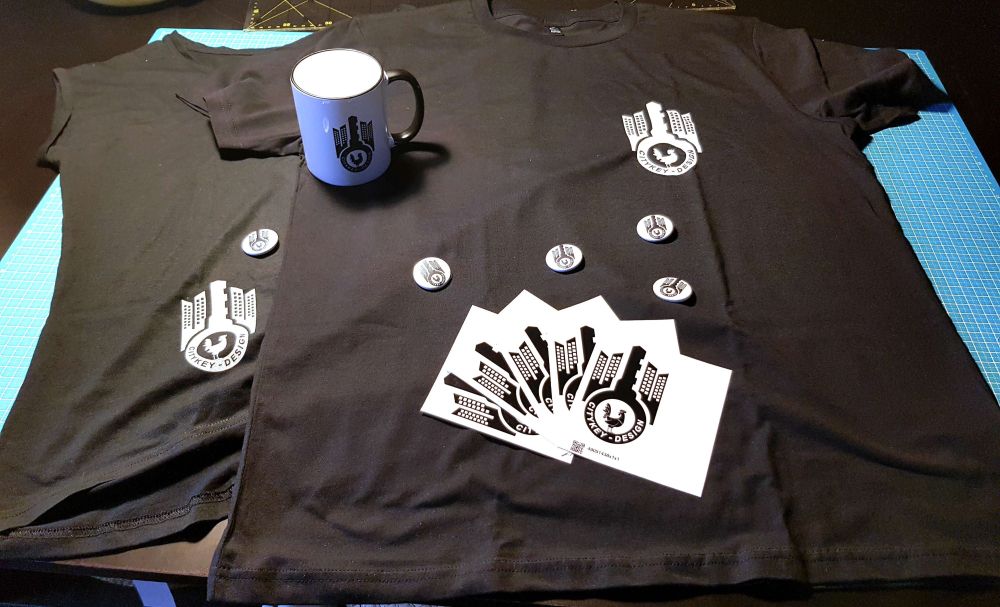 a picture of 2 black T-Shirts, a black and white mug, 5 buttons and 5 stickers all with the Citykey-Design-Logo.