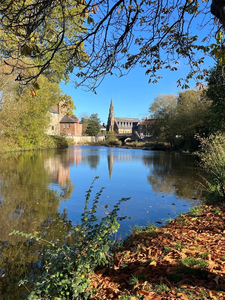 Photo looking downriver towards the Chantry, Telford Bridge and St George’s Church in Morpeth with gorgeous blue sky and autumn foliage framing the reflections in the water. 