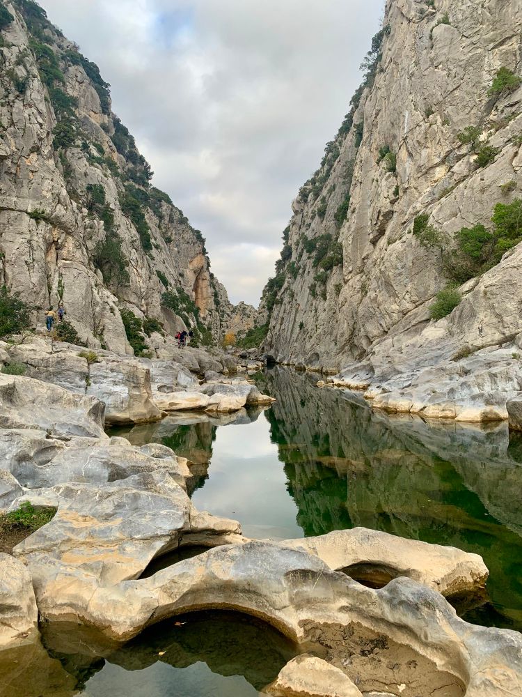 Gorges situées au pied de la grotte de Tautavel (66). Photo prise au niveau de la rivière entre deux falaises sous un ciel plutôt gris et nuageux. La falaise de droite se reflète dans l’eau comme une ligne de fuite. Au premier plan, la roche est usée par l’eau et forme une bassine circulaire.
