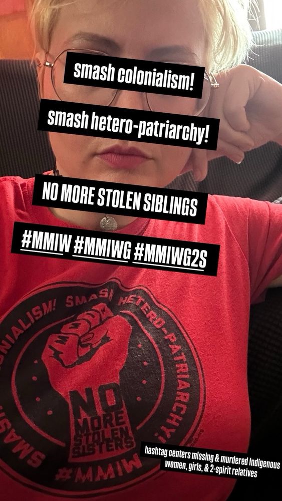image is of a light-complexioned bleach-blonde person wearing glasses and a red t-shirt with a design printed in black ink.  it is an upraised fist with M M I W tattooed on the knuckles.  wraparound text says SMASH CAPITALISM!  SMASH HETERO-PATRIARCHY!  NO MORE STOLEN SISTERS.  a website is included in very small font, www. Indigenous Action .org 