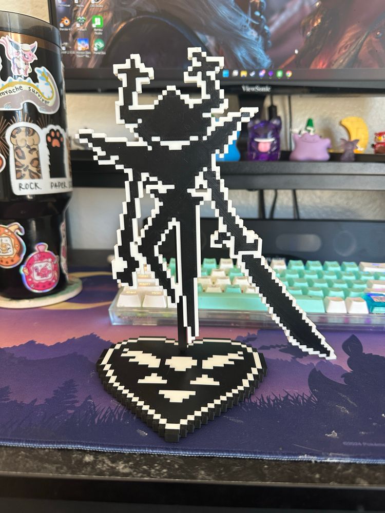Photo of an 8-bit styled, black & white standee of the Roaring Knight from Deltarune. She stands there menacingly, sword held out to the side and smiling. The base of the standee is of the Deltarune symbol. There is a black Stanley, with various stickers on it, to the left of the standee and in the background- a monitor can be seen and various trinkets, mostly Pokemon, on a raised shelf sit behind the standee. 