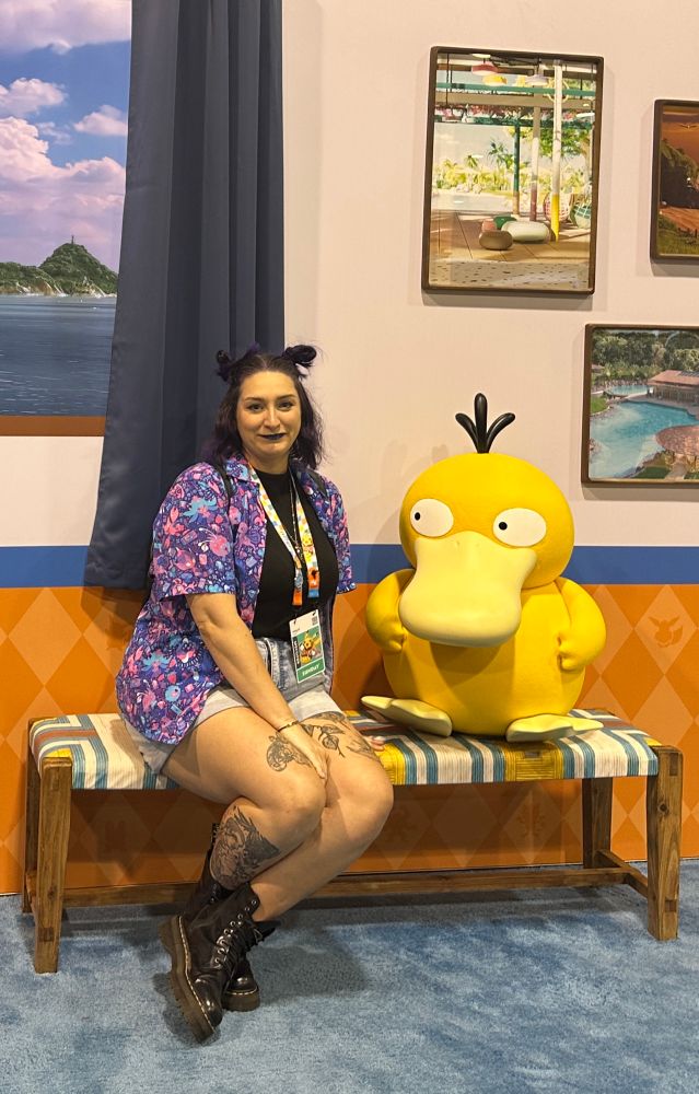 Photo of Ab smirking at the camera while sitting on a bench next to a Psyduck who stares at them a bit warily. The backdrop behind them depicts scenes of the island where the show Pokemon Concierge takes place. 