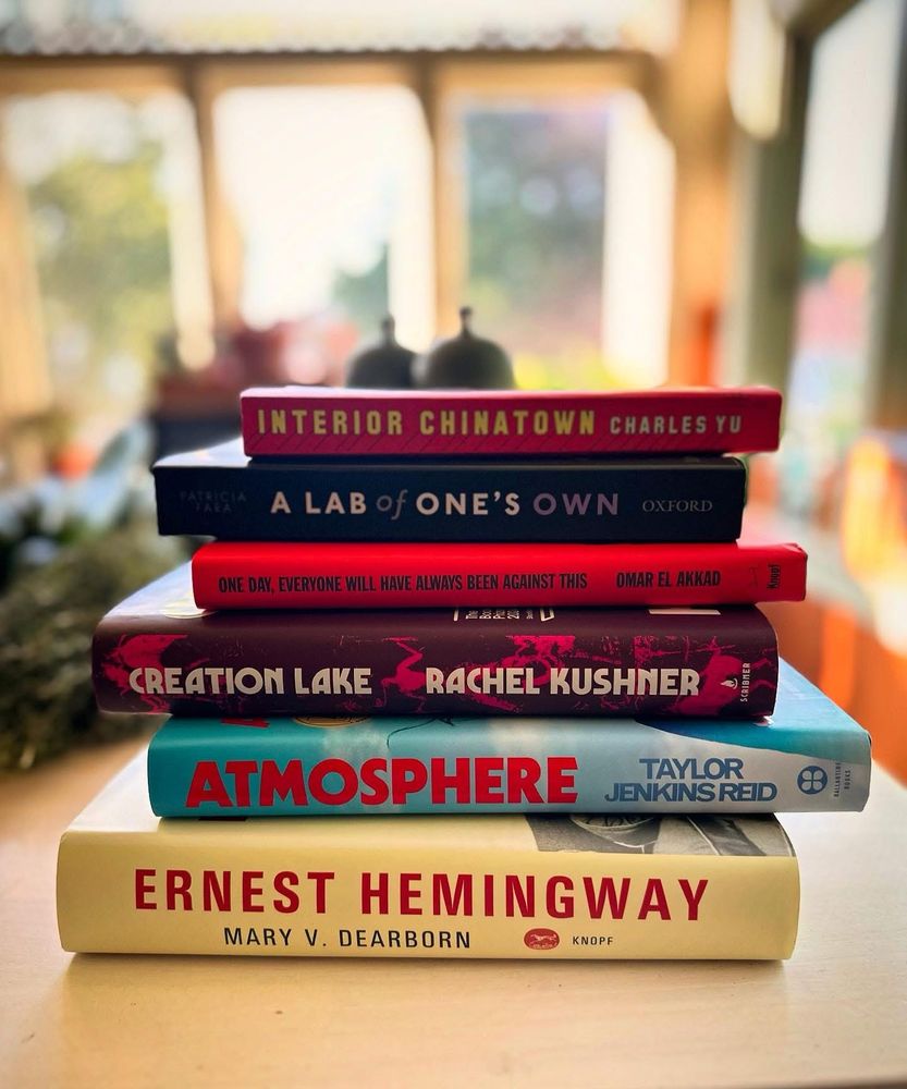 A stack of books on a white counter in front of a sunny window: Earnest Himingway by Mary Dearborn, Atmosphere by Taylor Jenkins Reid, Creation Lake by Rachel Kushner, One Day Everyone Will Have Always Been Against This by Omar El Akkad, A Lab of One’a Own, and Interior Chinatown by Charles Yu.