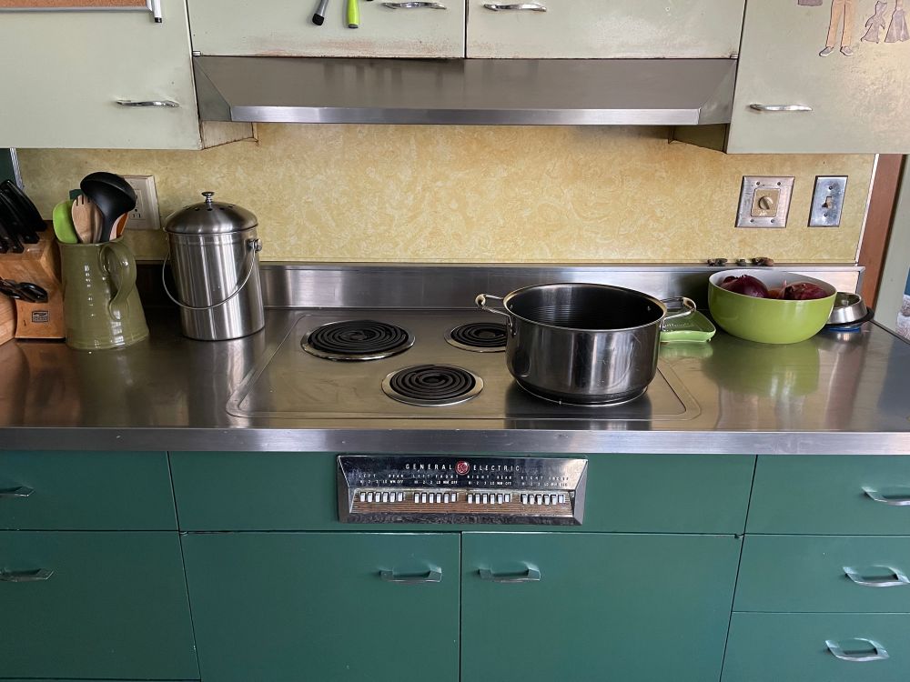 A modern photo of a 50’s style vintage kitchen counter; a steel counter over hunter green metal cabinets with a cooktop stove inset on the countertop. A large pot is set on top of the stove. The stove is General Electric and has push-button controls. 