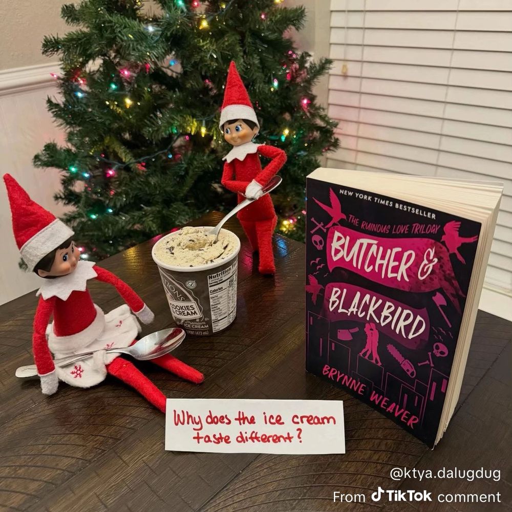 An Elf on the Shelf is scooping ice cream from a container while another Elf on the Shelf is sitting beside the ice cream container with a spoon on its lap. A note in front of them says “Why does the ice cream taste different?” Beside them is a book entitled “Butcher & Blackbird” by Brynne Weaver. 