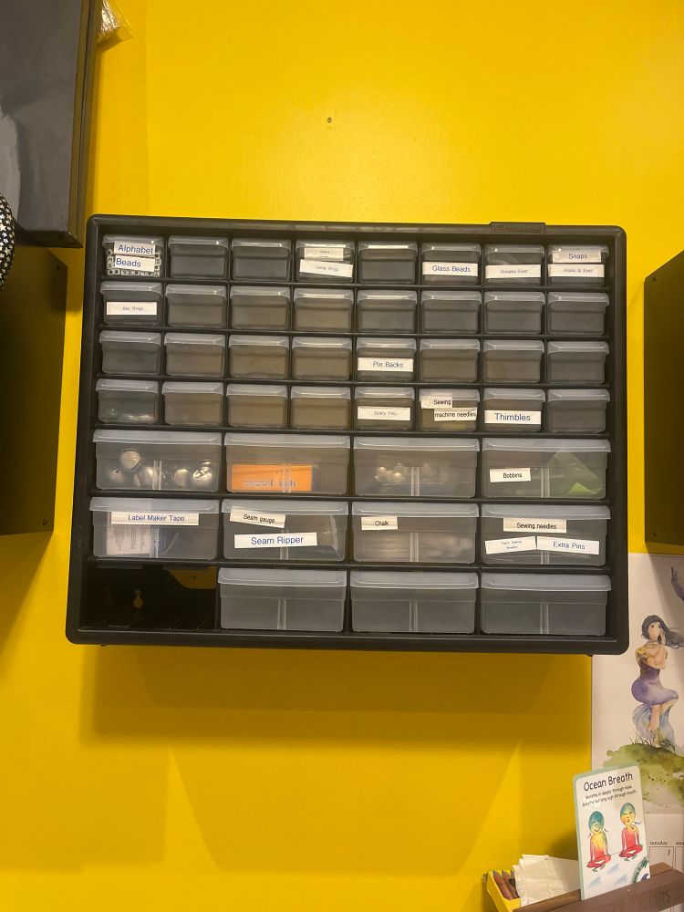 A plastic drawer storage unit hanging on a wall. Some of the drawers have labels on them and many of the drawers have contents that can be seen through the plastic drawer fronts.