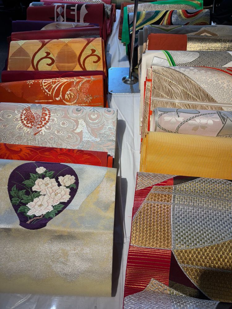 A table with heavily detailed and very beautifil kimono obi (belts). A whole range of colours and designs: flowets & geometric designs dominate. 