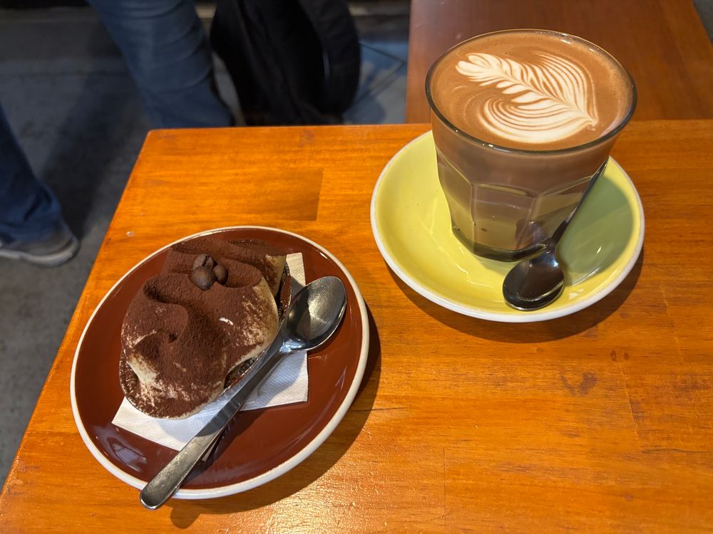 An Italian tiramisu sits upon a plate next to a mocha coffee. You can see the delicious swirls of cream on top that are covered with cocoa powder. The coffee has a leaf type design on the crema. 