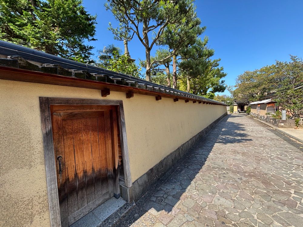 A long wall stretches into the distance. It is capped with black decorative tiles. A small wooden door is in the foreground. The street is paved with stone. The samurai district of Kanazawa. 