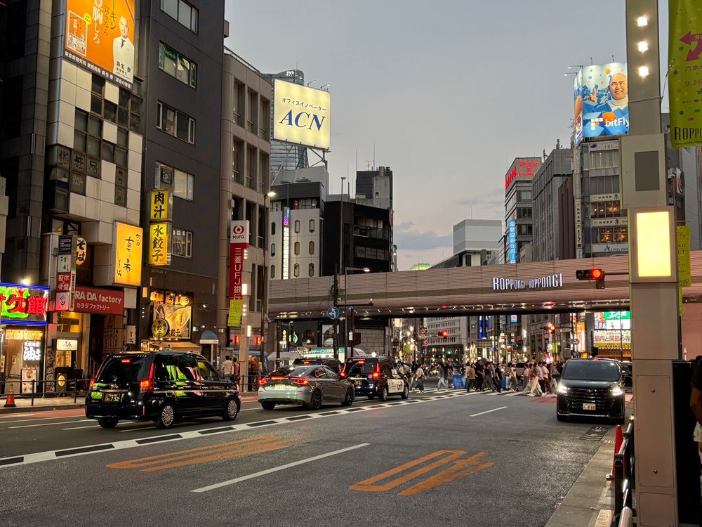The Roppongi intersection in Tokyo. A busy road with cars & buildings and the Roppongi lit sign on the overpass. It is dusk. Clear skies. 