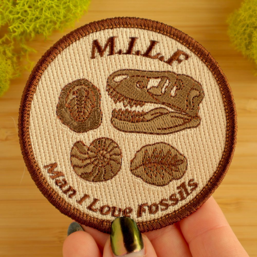 A photo of a circular iron on patch. It is a light tan brown with darker brown embroidered fossils of various types (a t-Rex skull, a trilobite, an ammonite, and a leaf). It has arching text above that reads M.I.L.F. Arching text below reads “Man I Love Fossils). 
