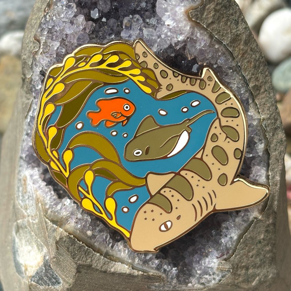 A photo of a large enamel pin featuring a leopard shark, kelp, garibaldi, and a bat ray. The pin is gold plated and has a dark blue ocean background. 