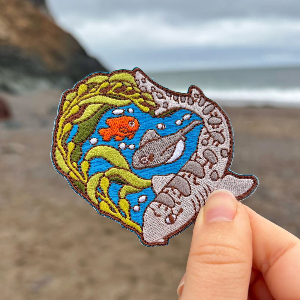 A photo of my hand holding an embroidered iron-on patch featuring a leopard shark, kelp, a bat ray, and garibaldi. The patch is being held in front of a beach and ocean background.