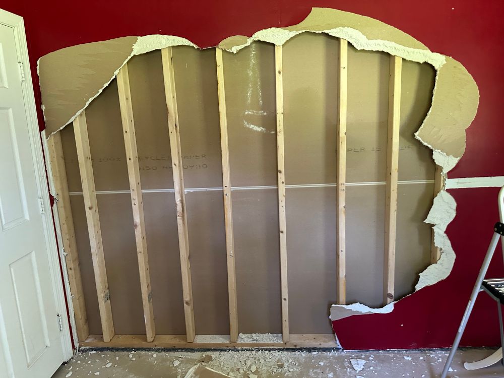 A partially demolished wall with red paint and exposed wooden studs. The drywall paper is ripped up along the edges 