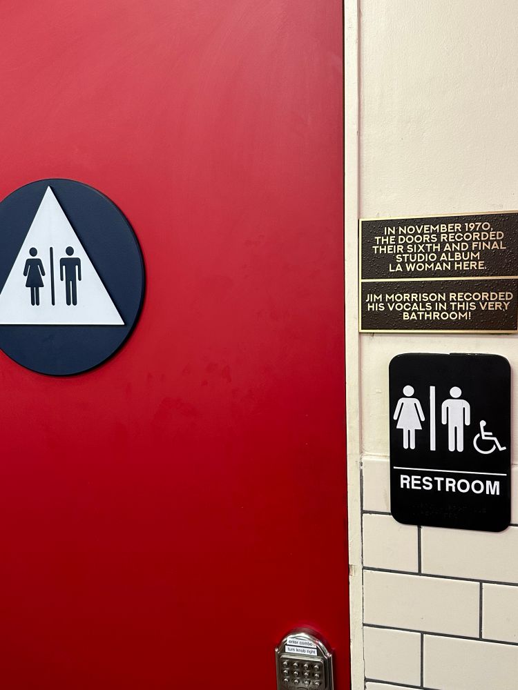 A door to a restroom that has a sign posted next to it that states “In November 1970 The Doors recorded their sixth and final album, LA Woman here. Jim Morrison recorded the vocals in this very bathroom”.