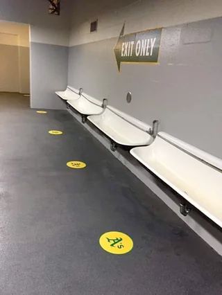 A men's restroom at the Oakland Coliseum with Oakland A's logos for safe distancing standing points in front of a row of four urinal troughs.