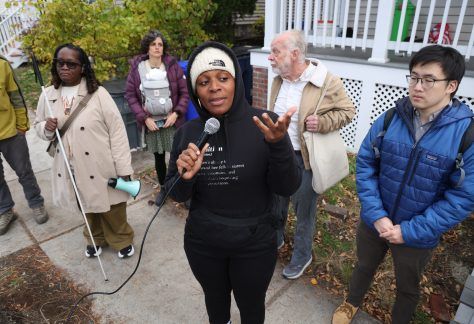 Group of people standing on sidewalk along Hyde Park Avenue. person in middle is using a microphone to describe why she discourages her son from riding his bike to school along / across Hyde Park Ave.