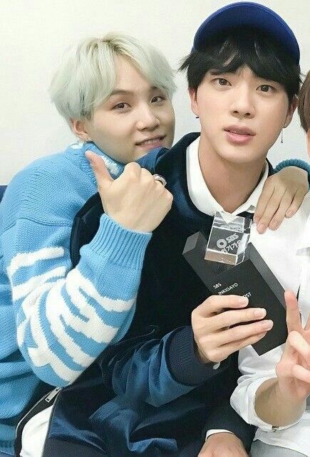 Blonde lightly mint Yoongi wearing sky blue sweater with white stripes and arm around Jin's shoulder with other hand giving thumbs up while Jin is holding award