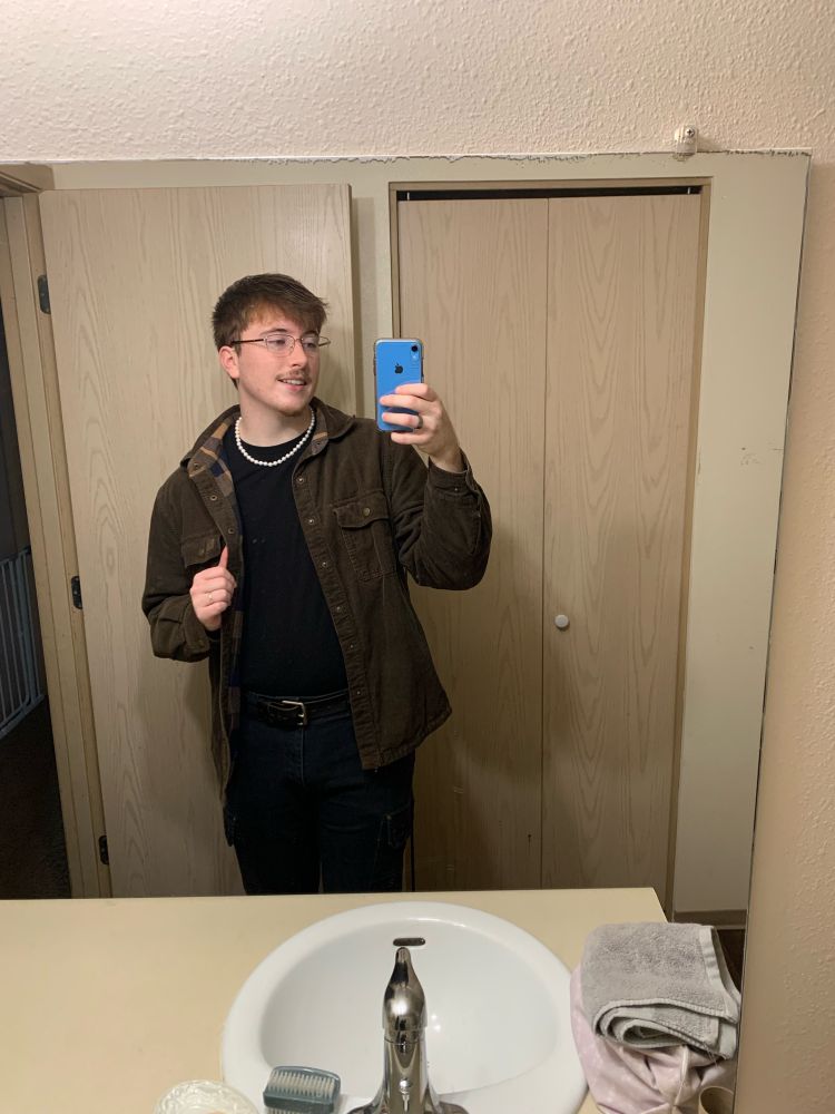 Brown jacket, black t shirt, white pearls, black work pants, brown belt, silver rings, and glasses on Brendan in a bathroom selfie