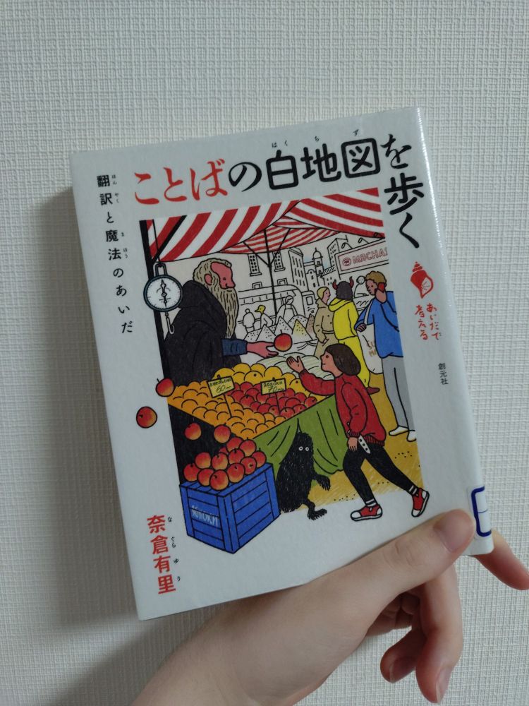 A Japanese nonfiction book being held against a wall covered in white, textured wallpaper. The pale hand of the poster (me) is visible holding the bottom of the book between thumb and pointer finger. The cover shows an illustration of a child receiving an apple from an older man with a long beard at a market. A small, black creature peeks out from under the stall.