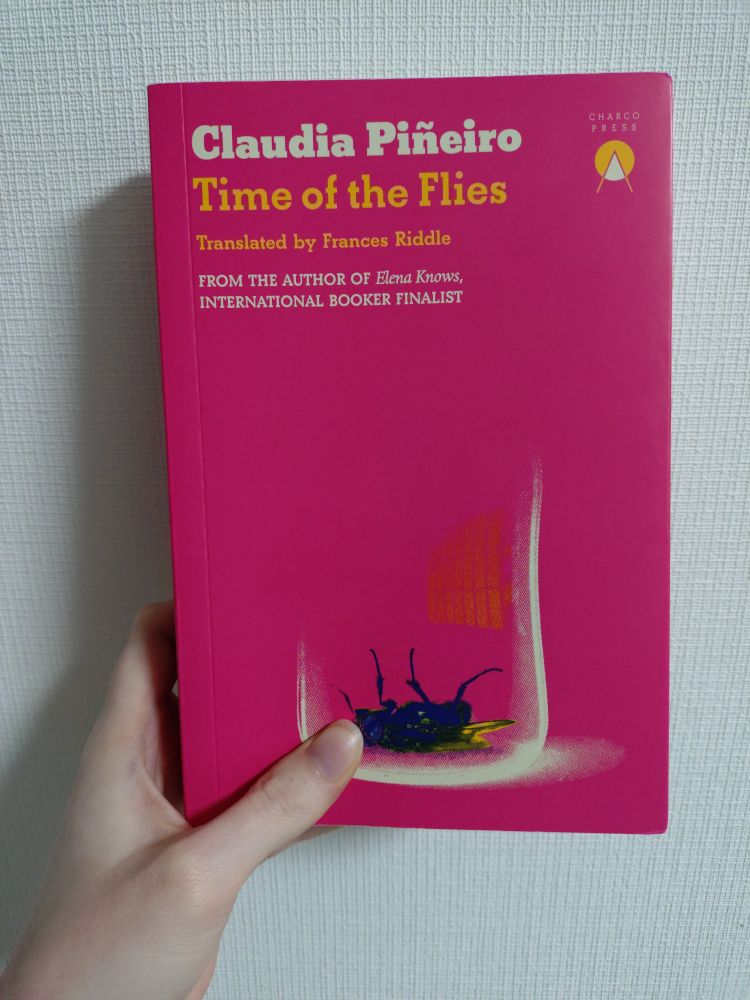 The novel Time of the Flies by Claudia Piñeiro (tr. Frances Riddle) being held up again a wall with white textured wallpaper. It is held from the lower left hand corner in order to avoid obstructing the cover. It is a bold pink with the title, author name, and translator name in a deep yellow. Underneath indicates that a previous book by the author, Elena Knows, was a finalist for the International Booker Prize when it came out. Beyond that there is a simple illustration of a fly flipped upside down. The silhouette of a glass can be seen over top of it.