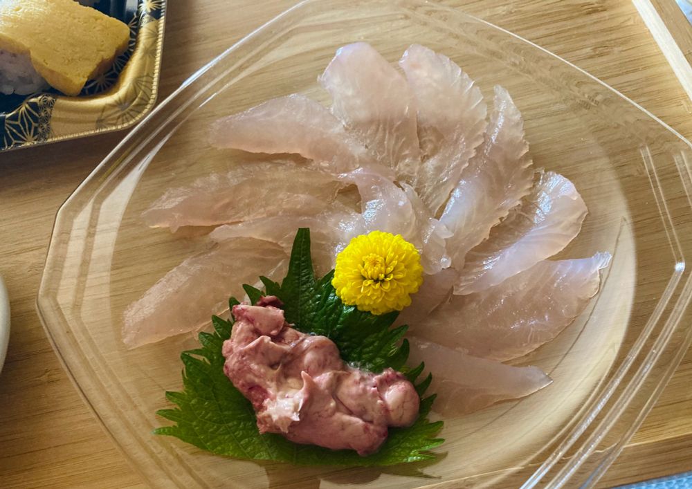 カワハギの刺身の写真。正方形の透明なパックに薄造りの、少しピンク色も混じった白身がぐるりと円を描くように盛られており、中心に食用菊、手前に、大葉に乗った肝臓が添えられている。