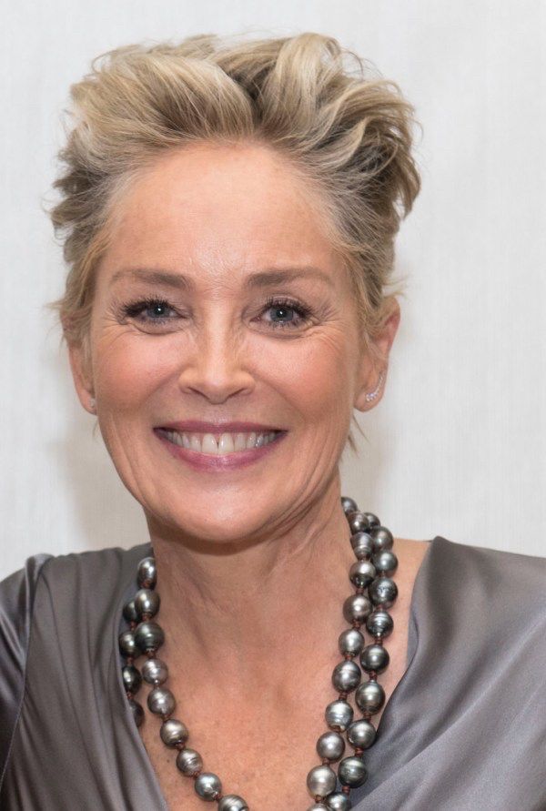 A smiling portrait of Sharon Stone, a renowned actress, with short blonde hair, wearing a black jacket with a high collar, captured against a neutral white background.