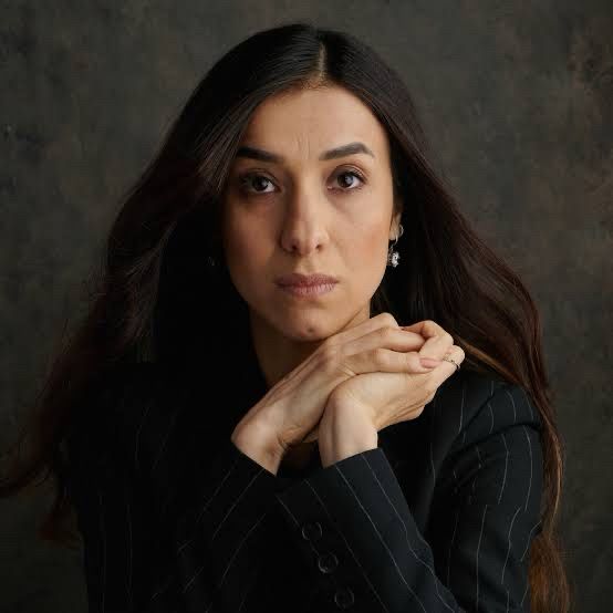 A portrait of Nadia Murad, a Nobel Peace Prize laureate, with long dark hair wearing a black pinstriped suit, gazing directly at the camera with her hands clasped under her chin against a dark backdrop.
	