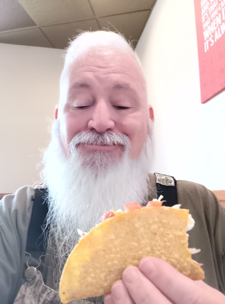 photo of an old fellow in camo coveralls holding a Taco that has a bite taken from it.