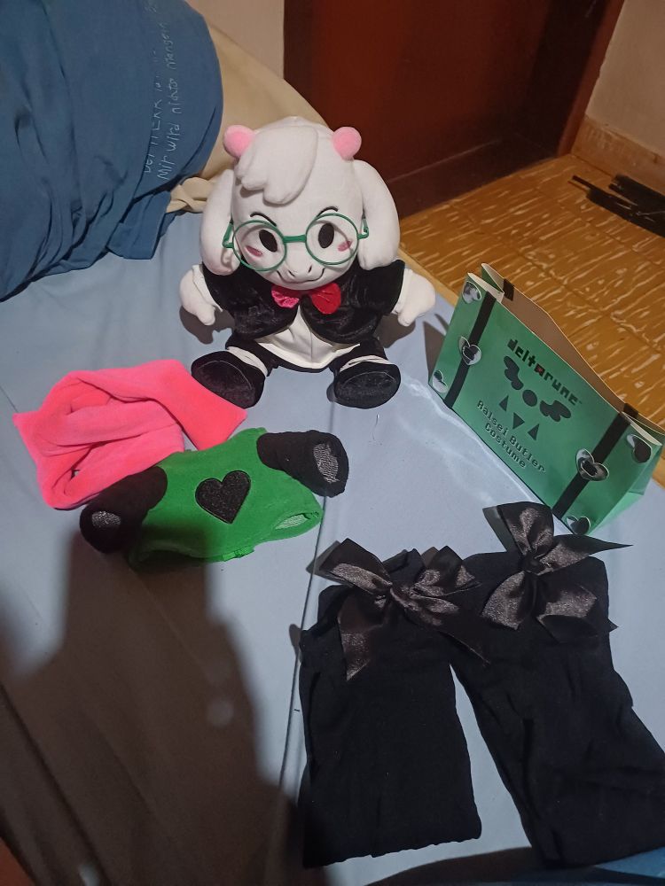 A Ralsei plush with a butler costume on sitting on a bed, with his original clothing just Infront of him. To the side of him there's a Deltarune branded bag, with thigh highs underneath said bag.