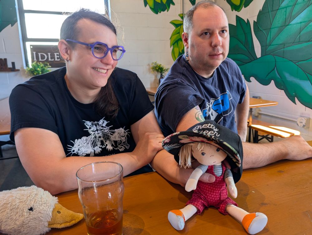 Ellie Lou and Brian sitting at a table at Idle Hands, behind Ellie Lou's doll Ollie, who is wearing Brian's "Pretty Queer" comically-oversized-for-a-doll baseball cap.