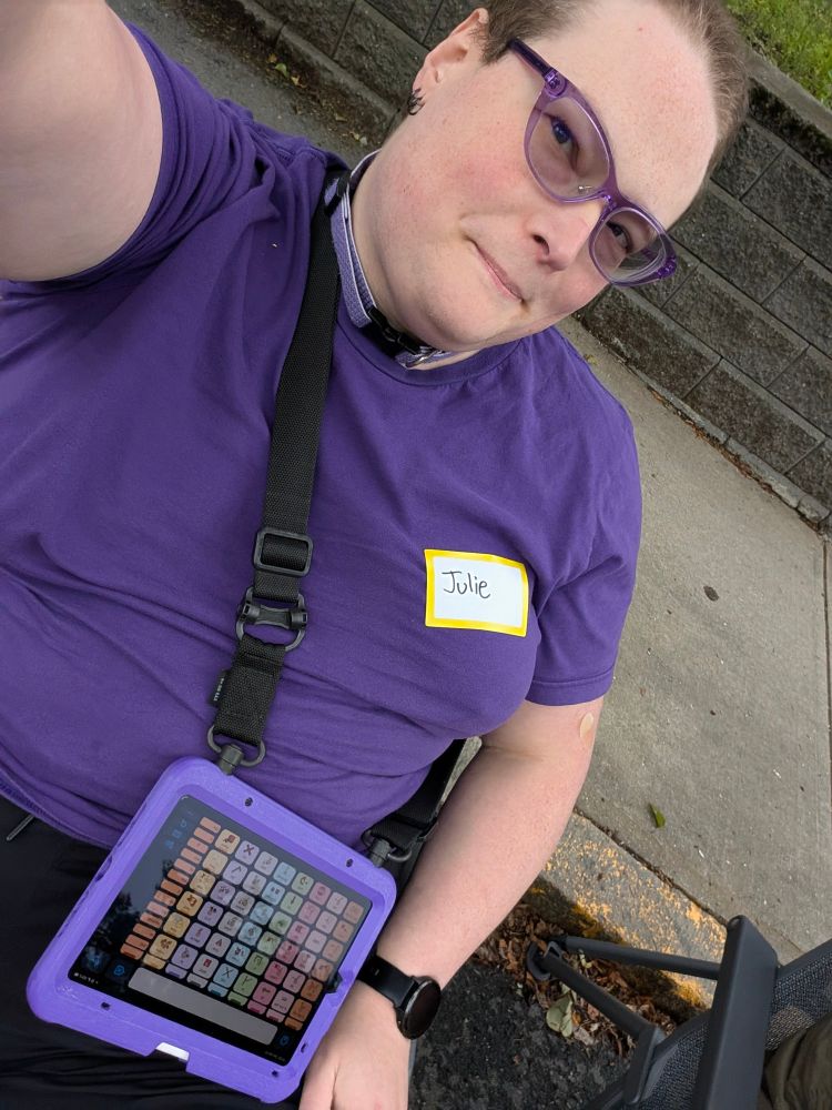 A fat white trans girl in translucent purple glasses, a light purple collar, a rich purple T-shirt, and black athletic shorts, wearing an iPad showing the Proloquo home screen, in a purple case on a shoulder strap.