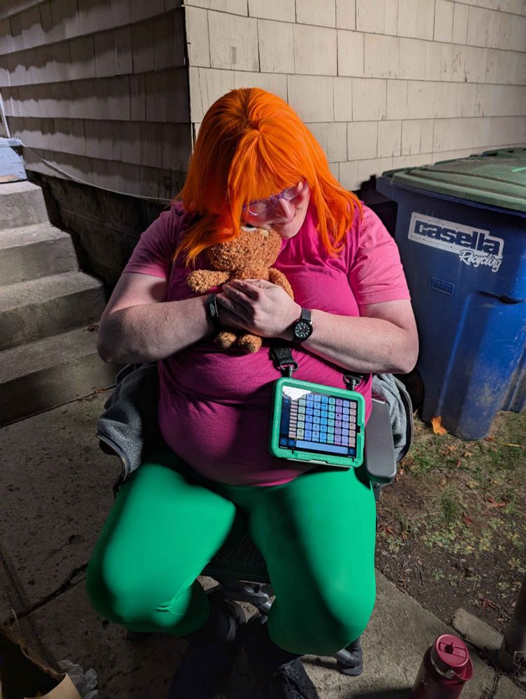 Me, sitting in a chair by my back steps, wearing a bright orange wig, "charity pink" T-shirt, magenta tunic, kelly green leggings, and black sneakers, holding Fluffster (a teddy bear plushie) and wearing my talker in a matching green case on a shoulder strap.