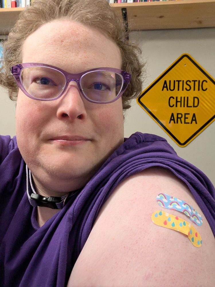 A white trans girl sporting two colorful Band-Aids on her left shoulder. She is wearing purple glasses, collar, and T-shirt and sitting in front of an "AUTISTIC CHILD AREA" road sign hanging on the wall beneath a shelf of books.