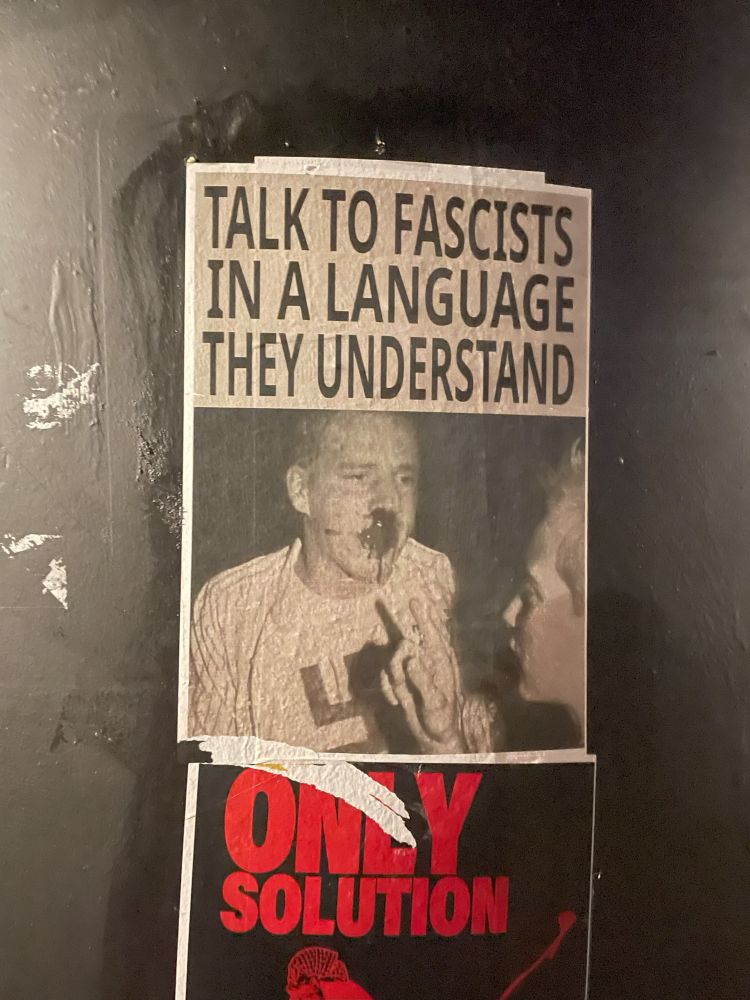 A poster with a black-and-white image of a skinhead man in a t-shirt with a swastika. another person is wagging a finger in the skinhead's face. the skinhead's nose and mouth are bloody. the caption reads "TALK TO FASCISTS IN A LANGUAGE THEY UNDERSTAND"