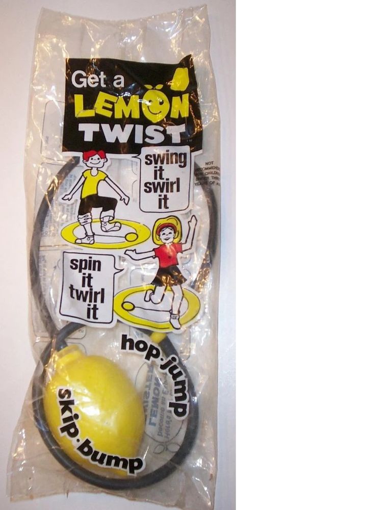 a Lemon Twist toy from the 1970s: a piece of grey plastic tubing with a yellow plastic lemon on one end and an ankle loop on the other. The toy is in a clear plastic bag with printed images of children playing with the toy, saying "swing it swirl it" "spin it twirl it" and additional printing reading "Get a ... LEMON TWIST" "hop jump" "skip bump".