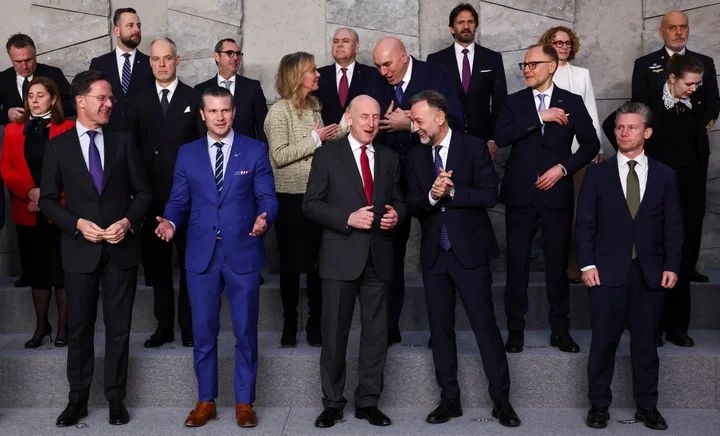 "family photo" of politicians at the NATO meeting today. Pete Hegseth is wearing an ultramarine suit with a Stars and Stripes pocket square and brown shoes. All the other guys are wearing black or dark blue suits and black shoes, and their pants have a break and hit at the top of the shoe, where Hegseth's pants have no break and hit above the ankle. He really stands out. 