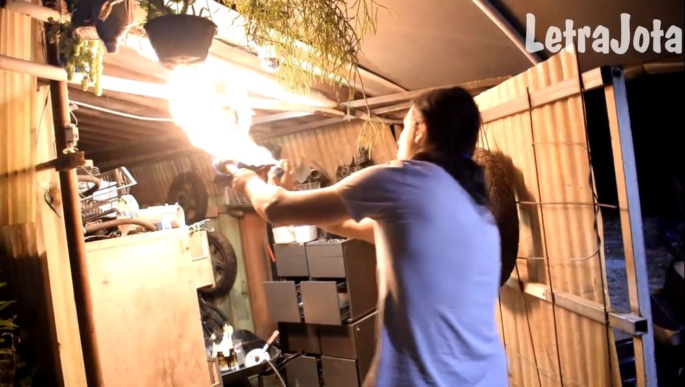 Momento do vídeo em que ele segura uma tocha em uma mão, e um desodorante na outra mão, projetando fogo dentro de um galpão de fundo de quintal. Se vê a panela de pressão ao fundo dentro de um carrinho-de-mão de obra.