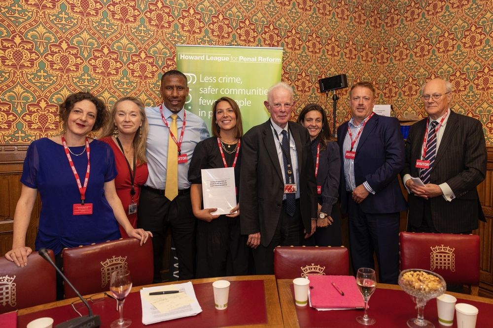 Eight people face the camera in front of a Howard League banner, in a committee room in the House of Lords. A woman with brown hair holds a copy of the report.