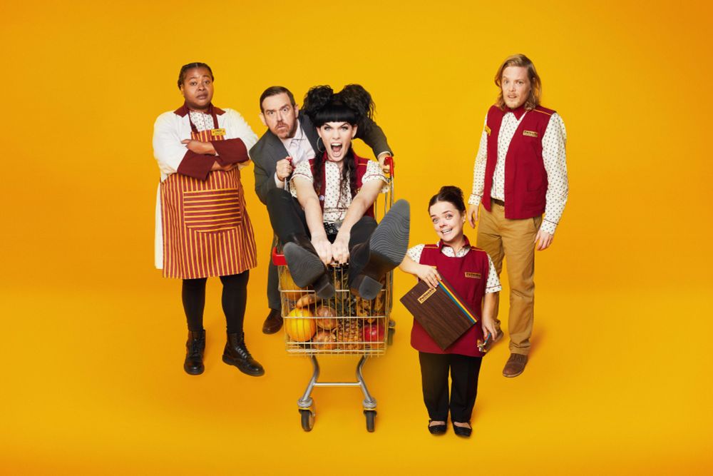 Key art for Transaction. Kayla Meikle, Francesca Mills and Thomas Gray stand around Nick Frost, who is pushing Jordan Gray in a shopping trolley. All are wearing supermarket uniforms against an orange background.