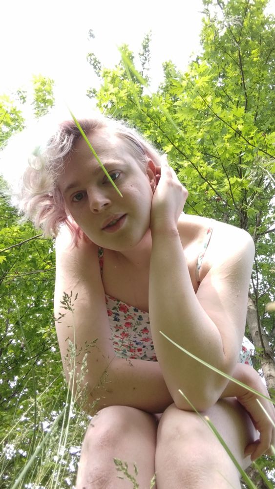 woman with short, slightly curly, multi-colored light-pink hair. she has deep blue eyes, and she's wearing a white patterned romper with blue and red flowers. she's posing outside, kneeling down to look at the camera placed in the grass, her hair falling down, one hand behind her ear (viewer right side), and her other hand across her knees (viewer left side). behind her overhead stretch the branches of a bright green tree, the sky glowing, casting her outline in a brilliant light