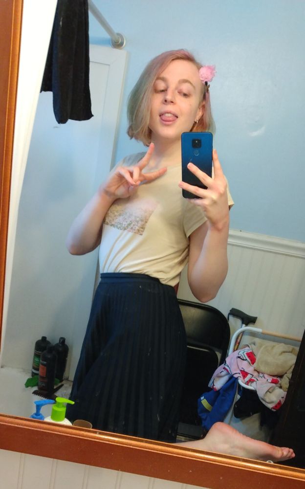 woman with short, multicolored pink/purple hair, wearing a light-colored t-shirt (with graphic of a pink flower field), long blue pleated skirt, and a small pink flower pinned in her hair (viewer's right side). she's posing in a bathroom mirror, body turned slightly towards the camera, with one leg up, one hand holding the phone, and her other hand in a peace sign