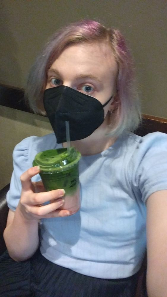woman with short, multi-colored pink hair, blue eyes, wearing a light-blue top, dark blue skirt, and a face mask. she's posing sitting down, looking up at the camera, and holding an iced matcha latte- it's multiple layered colors, with a dark red at the bottom, a milky pinkish red in the middle, fading to dark green on top