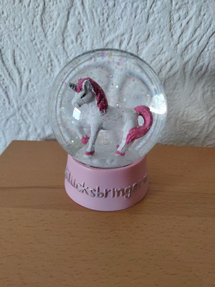 A snowglobe with a pink base, labeled "Glücksbringer" ("Lucky charm" in german). Inside the globe is the figure of a prancing white unicorn with pink hooves, mane and tail and a silver horn. The snow is made out of pink and iridescent flitter. 