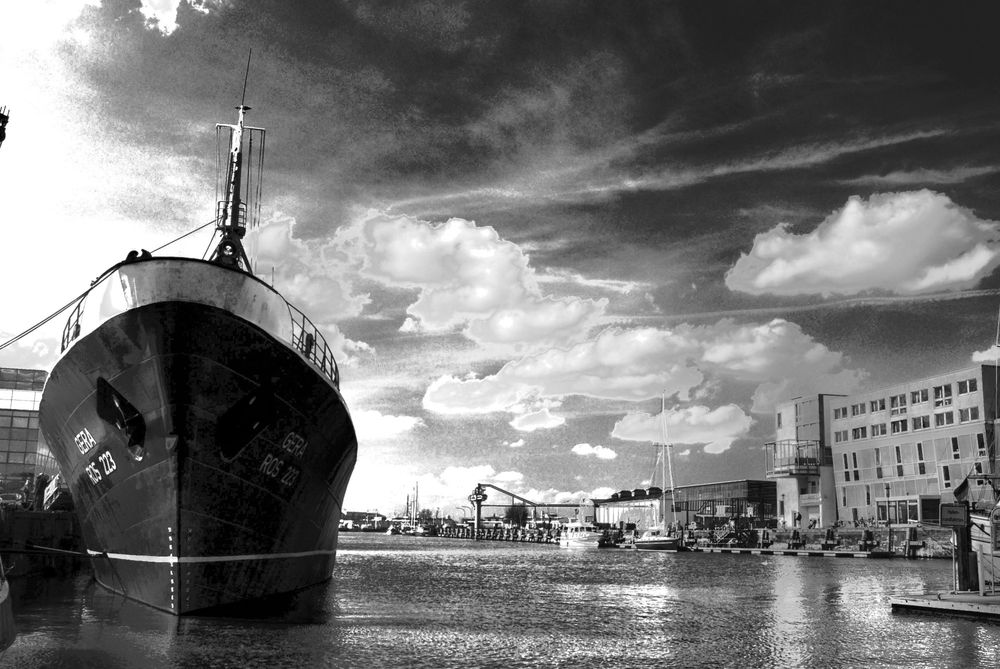 Links liegt ein altes Frachtschiff im Hafenbecken, im Hintergrund sind Hafenanlagen zu erkennen. 