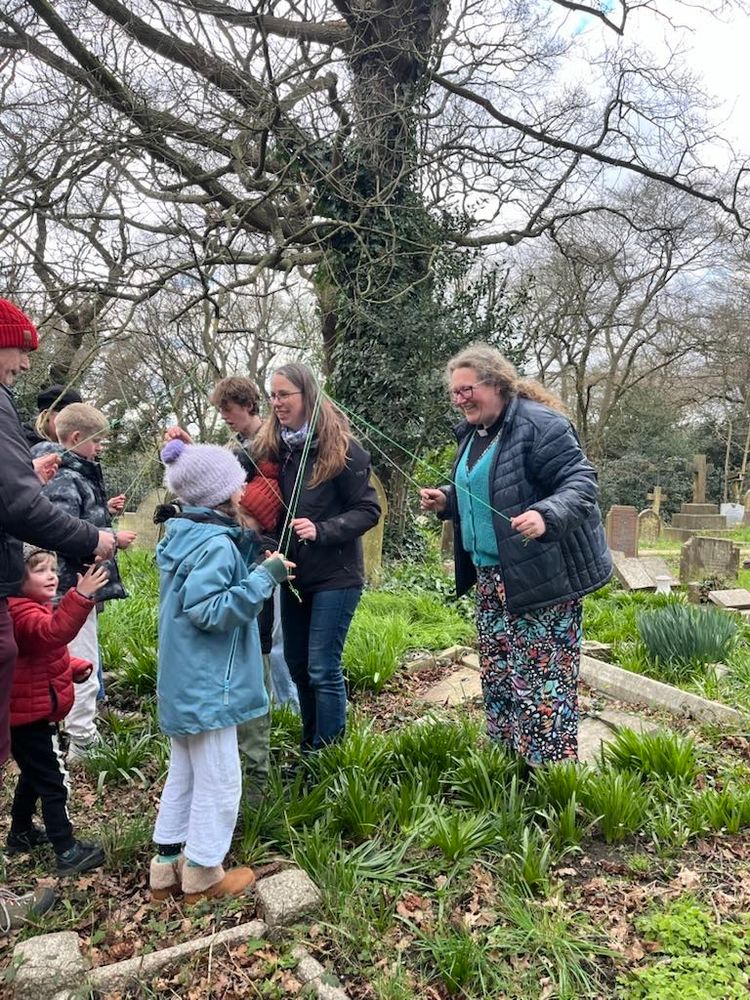 A group of mixed age people doing Nordic slinging braids under an oak tree in a churchyard.